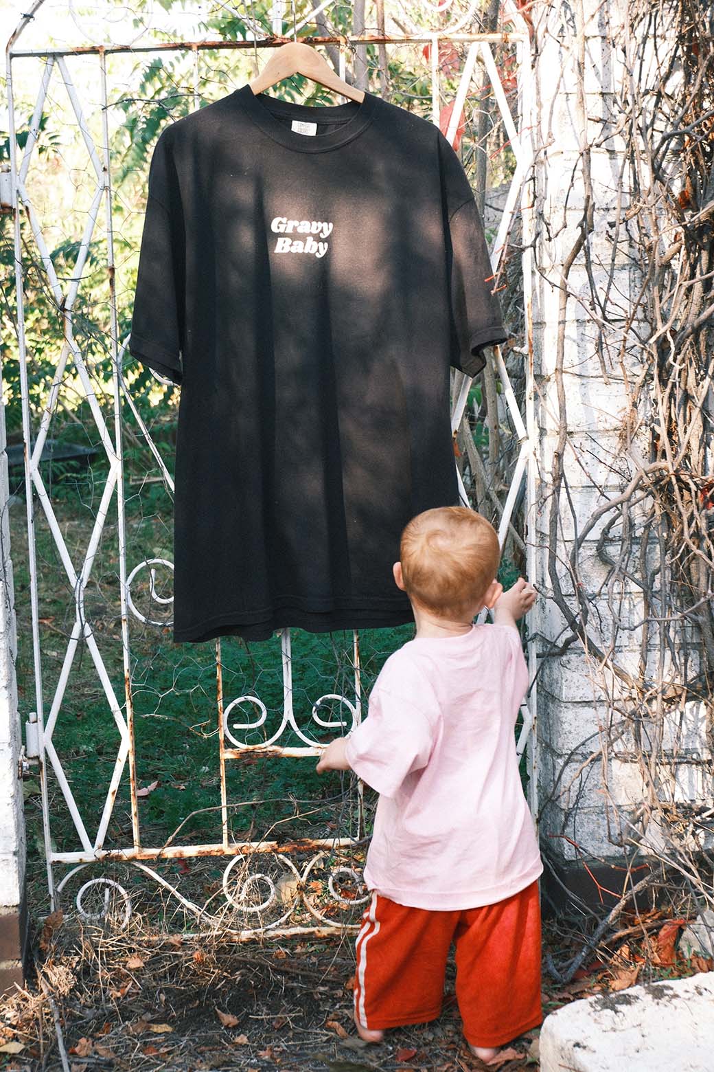 Black t-shirt with Gravy Baby text hanging on a metal gate, child in pink shirt and red shorts standing below.