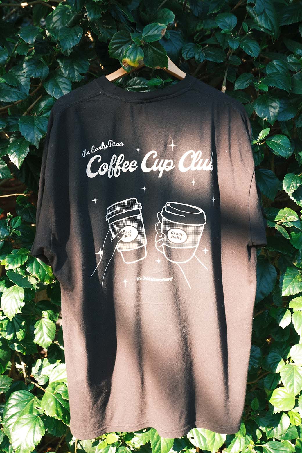 Black t-shirt with 'Coffee Cup Club' design on a hanger against a leafy background