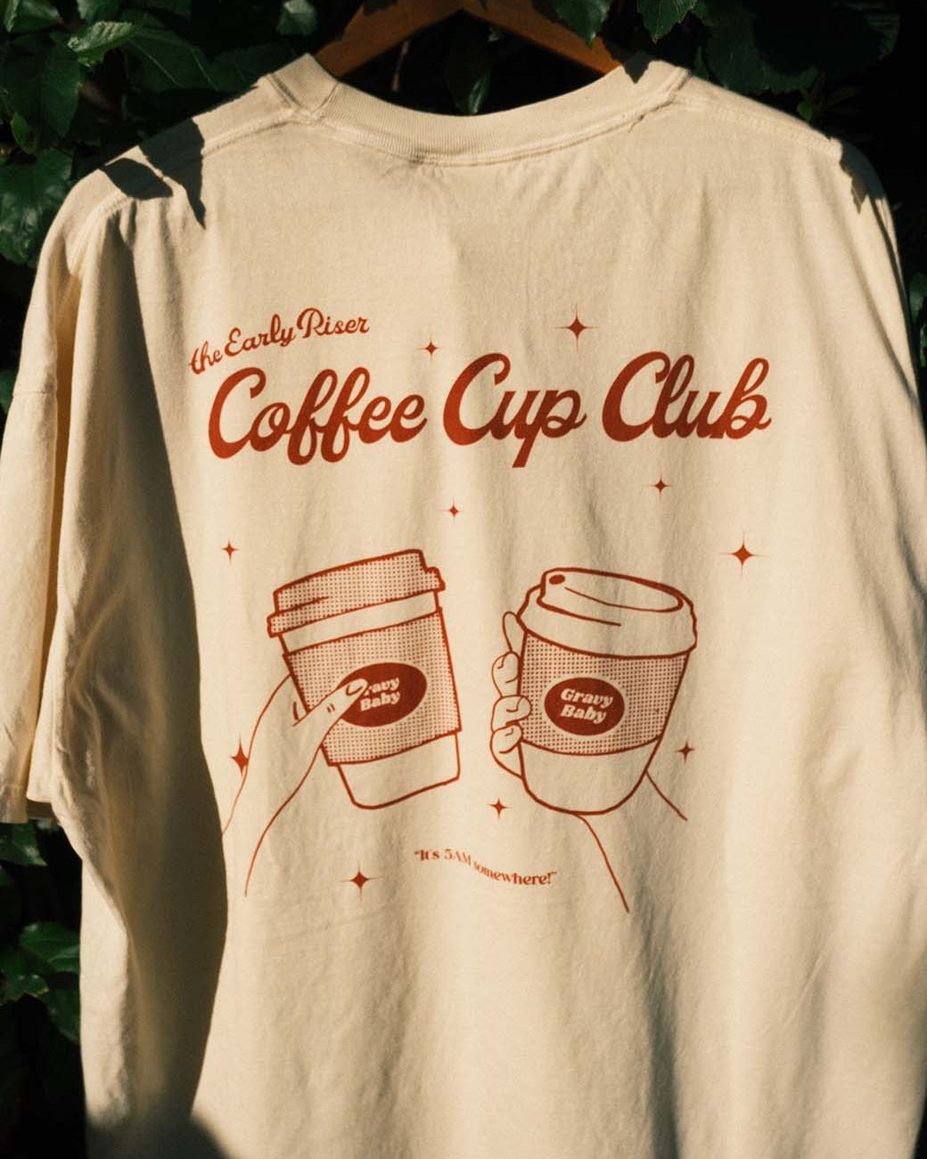 Beige t-shirt with 'Coffee Cup Club' design on a hanger against a leafy background