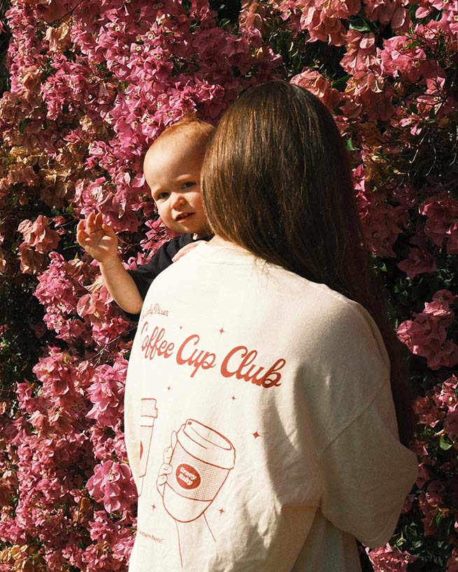 Woman holding a baby in front of pink flowers wearing a 'Coffee Cup Club' shirt.