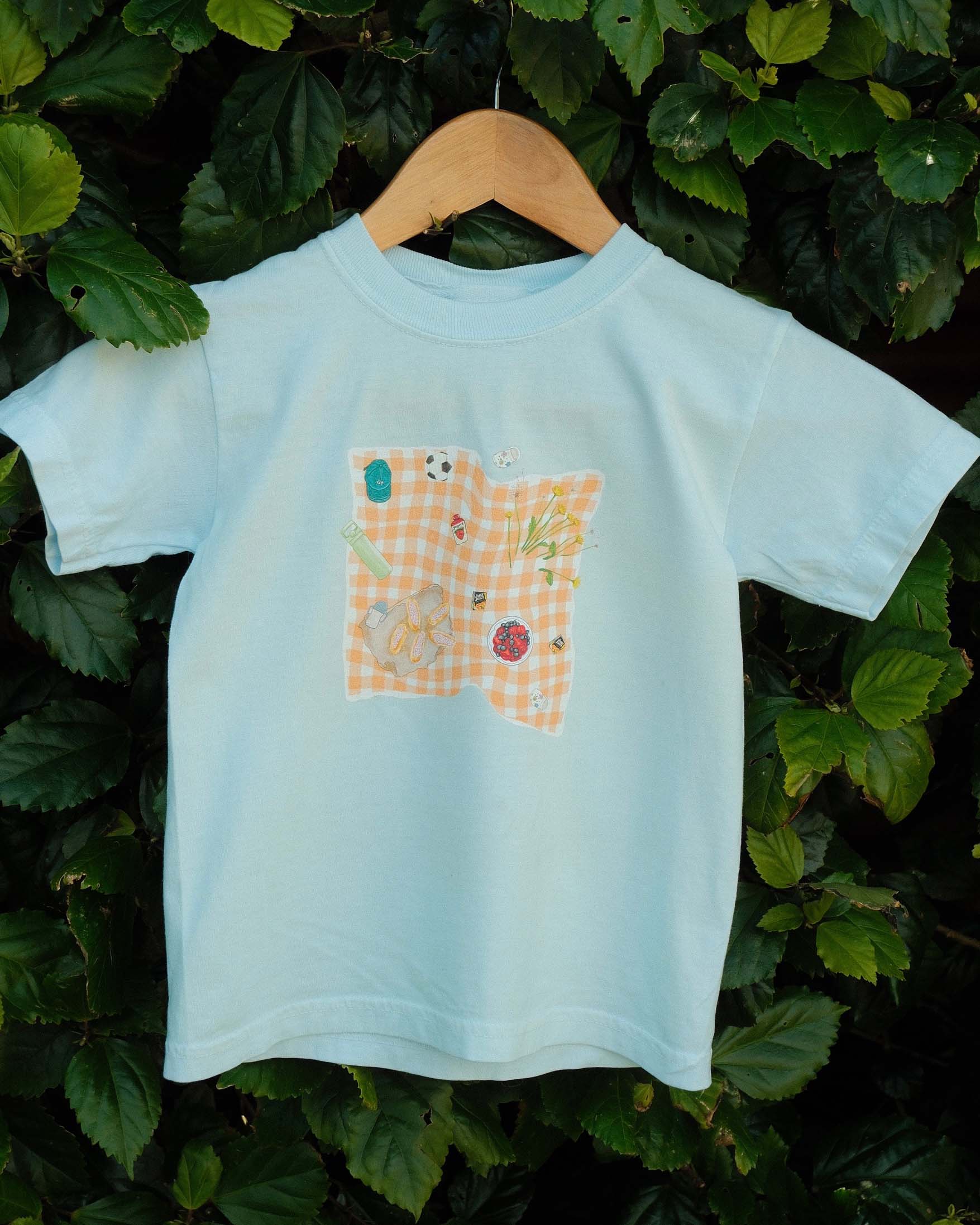 Light blue t-shirt with a picnic blanket design on a wooden hanger against a green leafy background