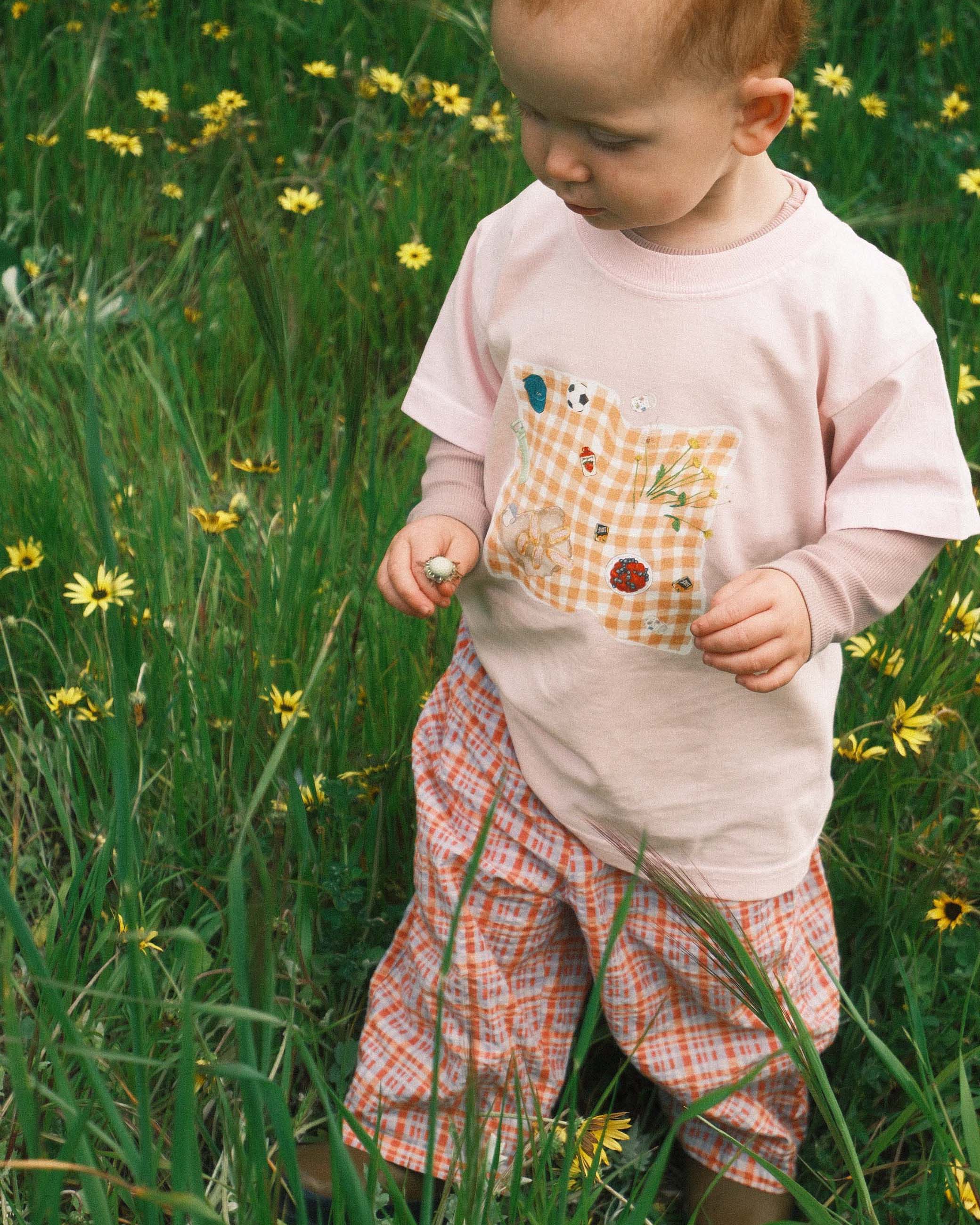 Child in a field of tall grass and wildflowers wearing a light pink shirt with a pocket design and plaid shorts.