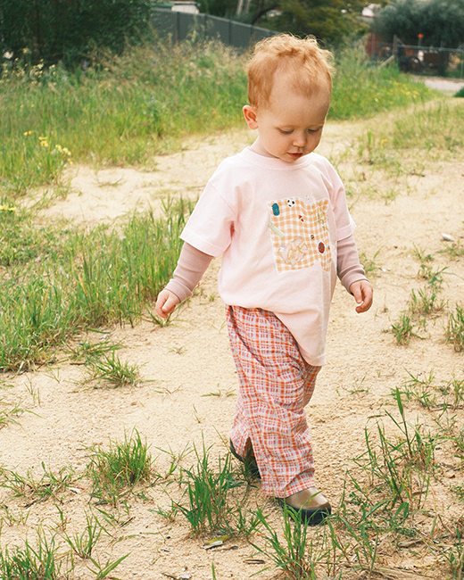 Child in a pink shirt with a patterned pocket and plaid pants standing on a dirt path with grass.
