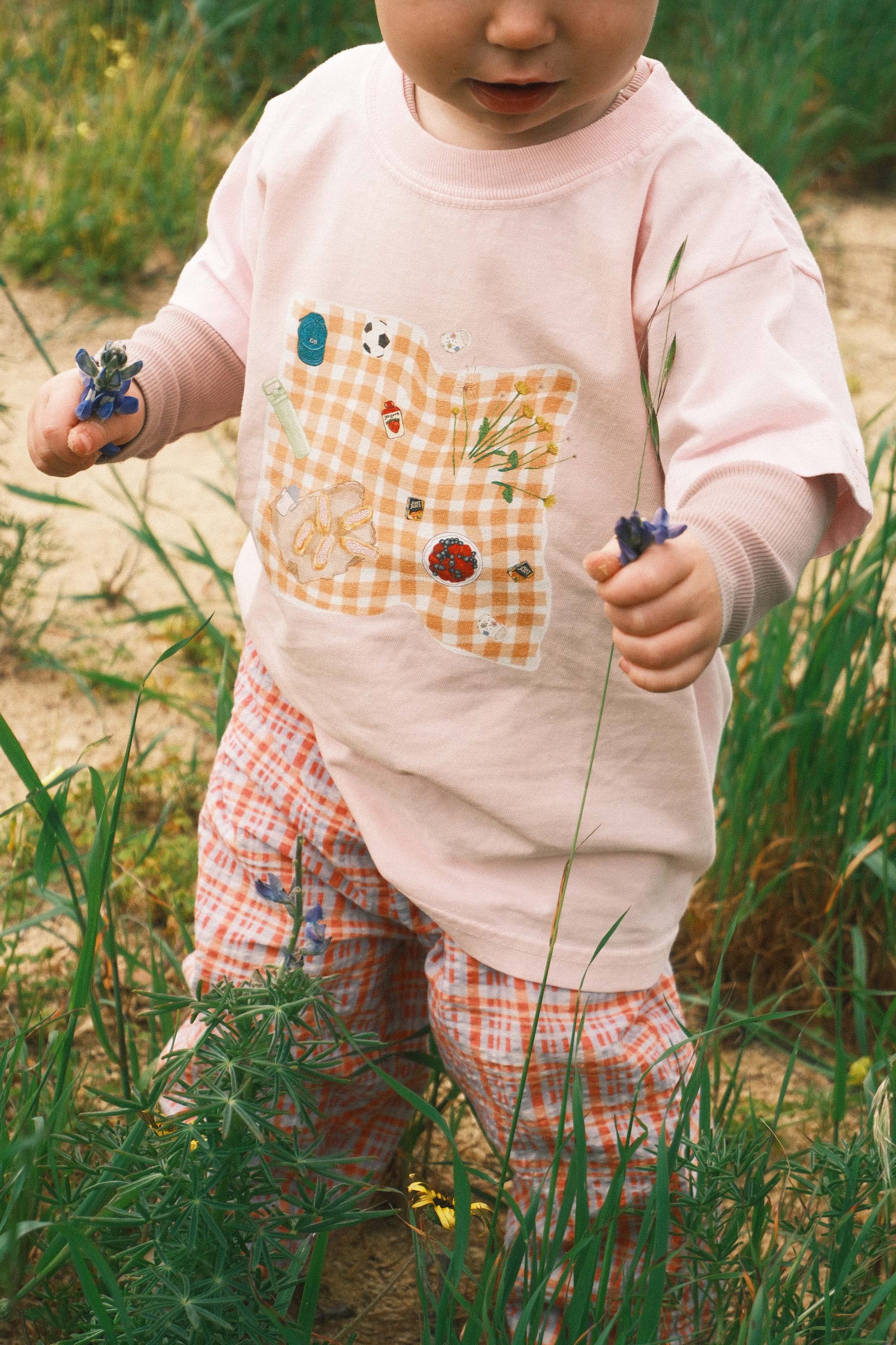 Child in a pink shirt with a checkered pattern and plaid pants standing in grass.