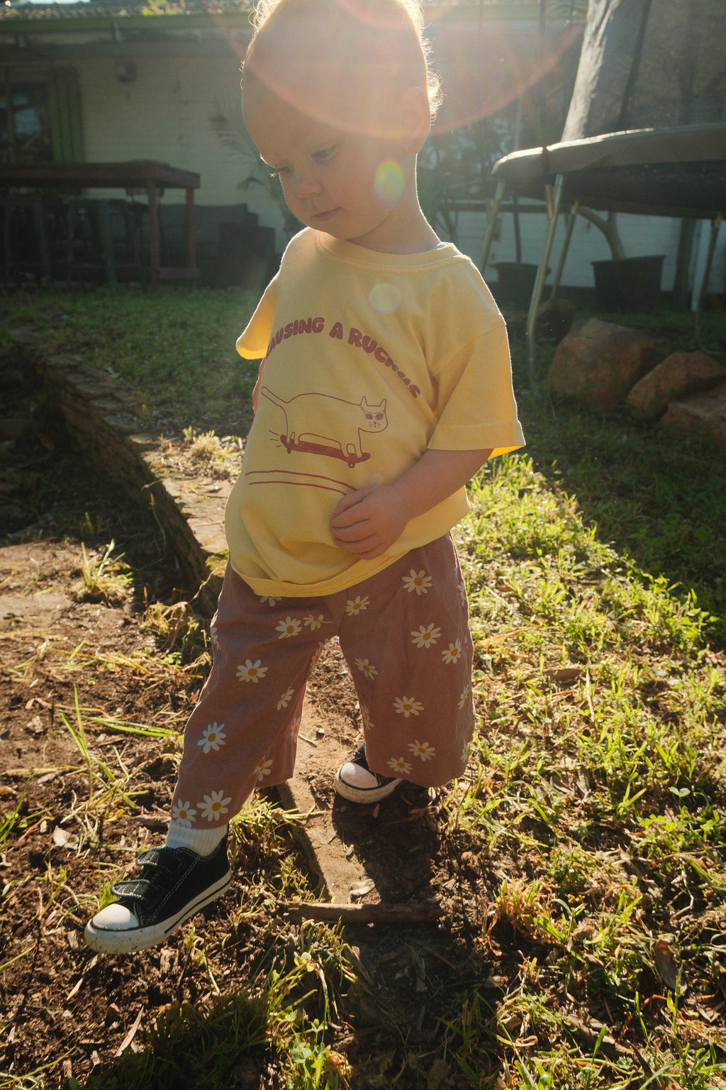 Child in a yellow shirt and patterned pants standing outdoors on a grassy area.