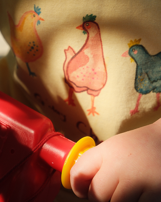 Child's hand holding a red toy with a yellow top against a fabric background with chicken prints.
