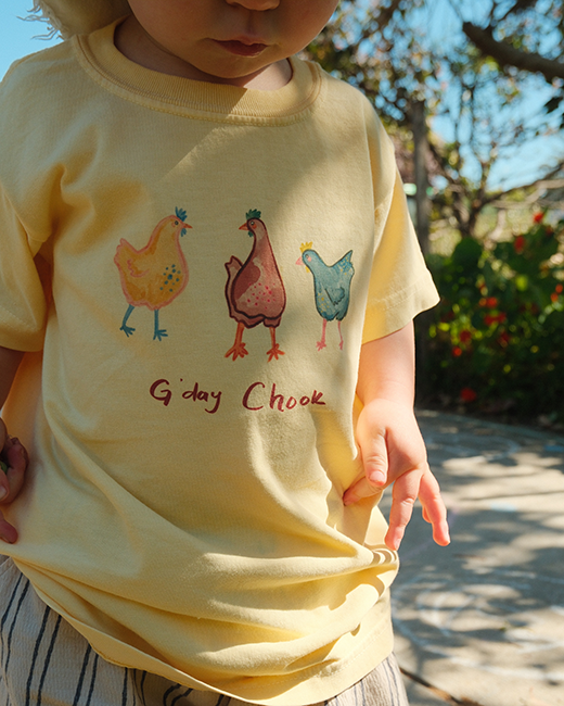 Child wearing a yellow t-shirt with cartoon chickens and 'G'day Chook' text outdoors.