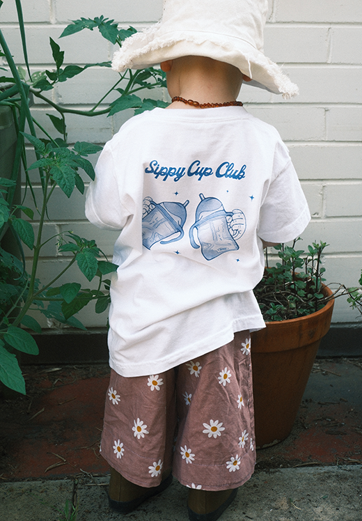 Child wearing a white t-shirt with 'Sippy Cup Club' design and floral pants, standing outdoors.