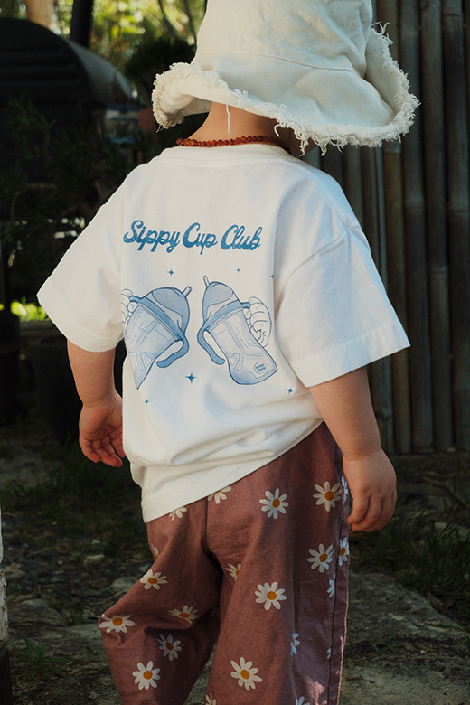 Child wearing a white t-shirt with 'Sippy Cup Club' design and floral pants outdoors.