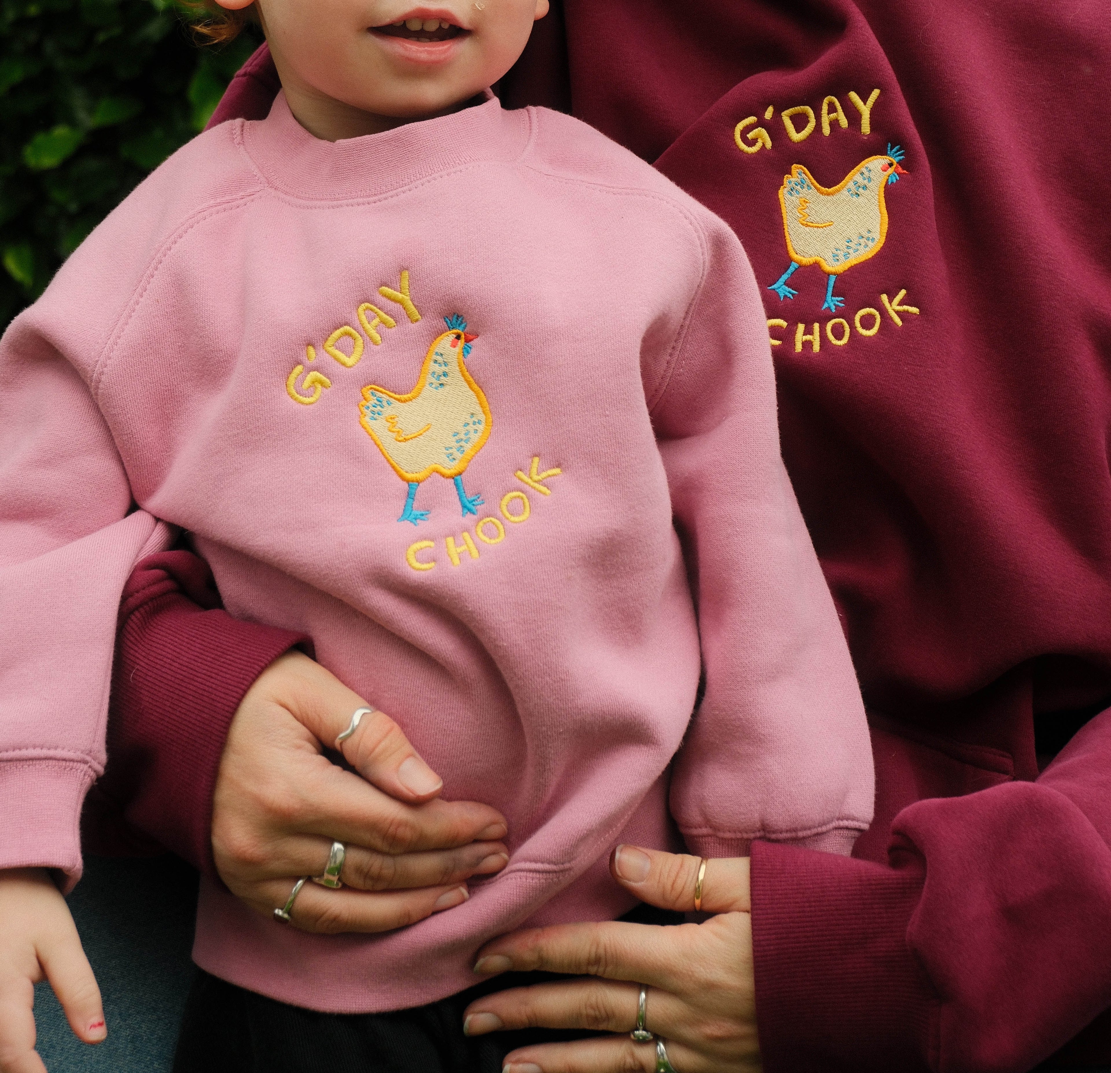 Child wearing a pink sweatshirt with 'G'DAY CHOOK' design, held by an adult in a matching maroon sweatshirt.