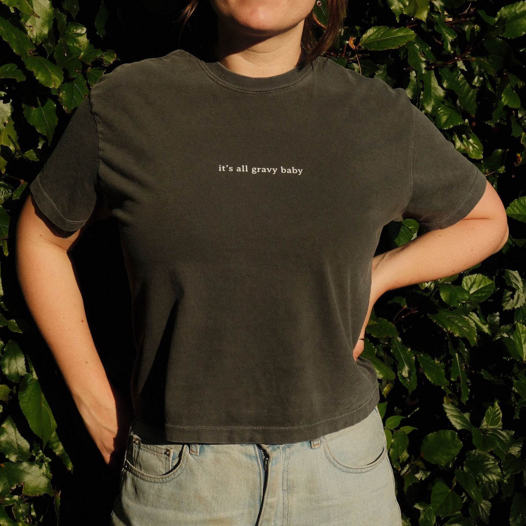 Person wearing a black t-shirt with 'it's all gravy baby' text in front of green foliage.