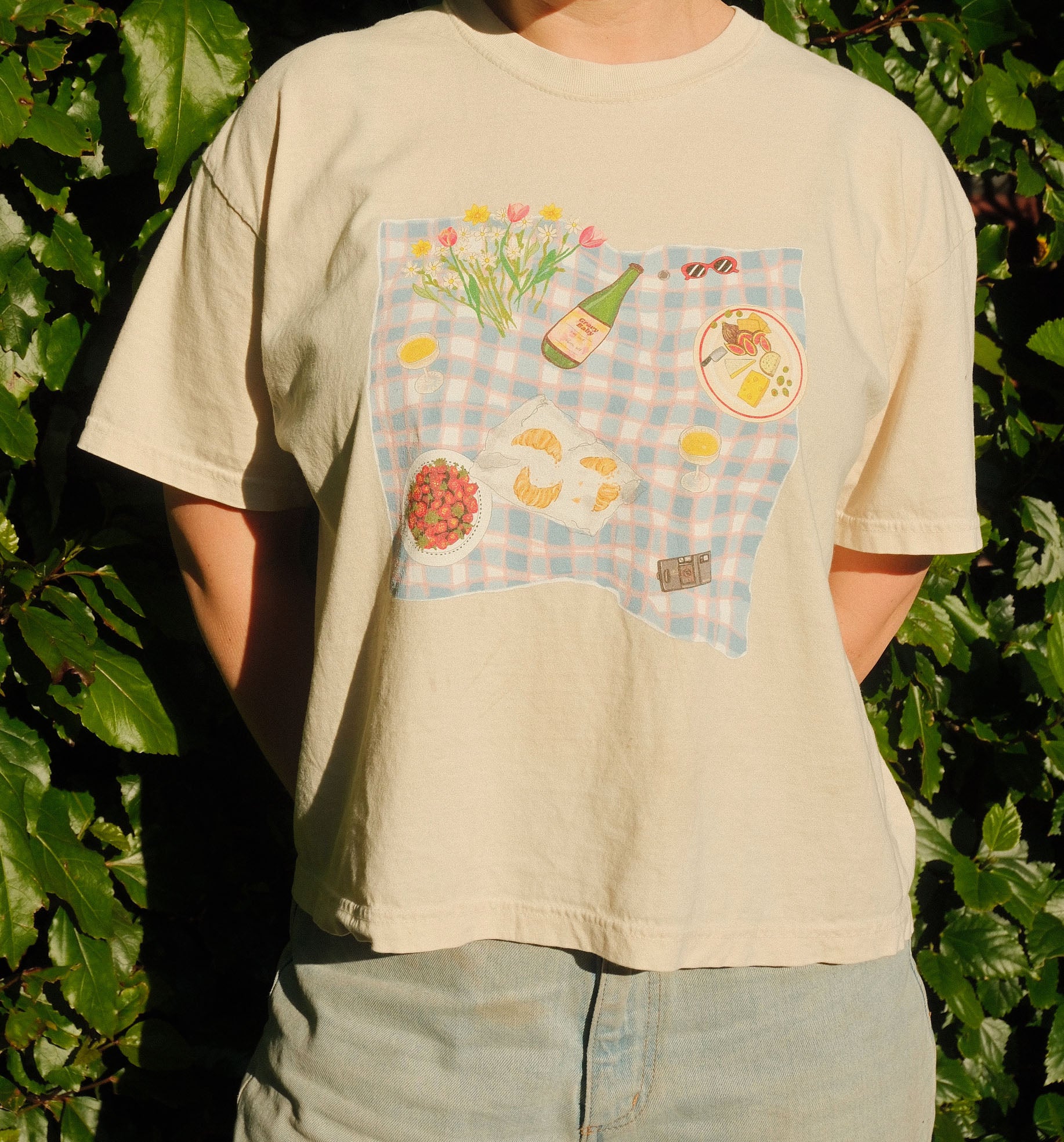 Person wearing a beige t-shirt with a colorful graphic design against a green leafy background